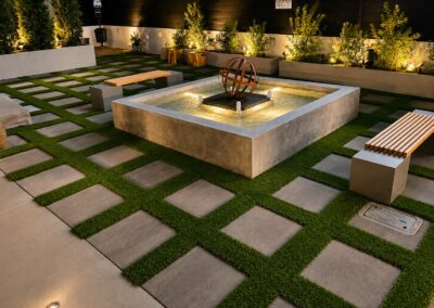 Modern outdoor lighting and hardscape design featuring illuminated water feature, paver walkways, and artificial turf landscape